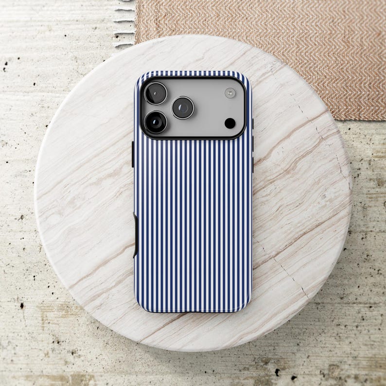 May include: A smartphone case featuring a vertical pinstripe pattern in navy blue and white. The case has a rectangular camera cutout at the top, revealing three circular camera lenses and a small flash component on the device underneath.