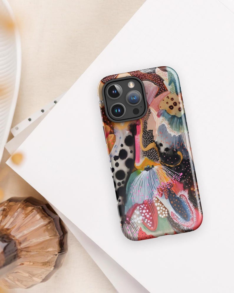 May include: A smartphone case featuring an abstract design with a mix of black, white, pink, blue, and tan colors. The pattern includes organic shapes, dotted textures, and fine line details across the entire surface of the rectangular cover.
