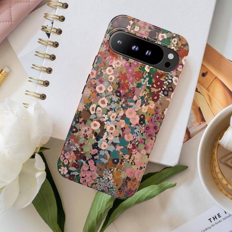 May include: A rectangular smartphone case featuring a dense, multi-colored floral pattern with shades of pink, green, brown, and blue. The case has a black camera bar cutout at the top, exposing the lenses of the device underneath the protective cover.