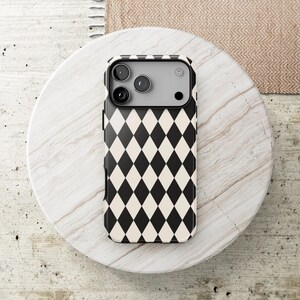 May include: A smartphone case featuring a repeating black and cream-colored diamond harlequin pattern. The case has a rectangular camera cutout at the top, revealing three circular camera lenses and a flash component on the device underneath.