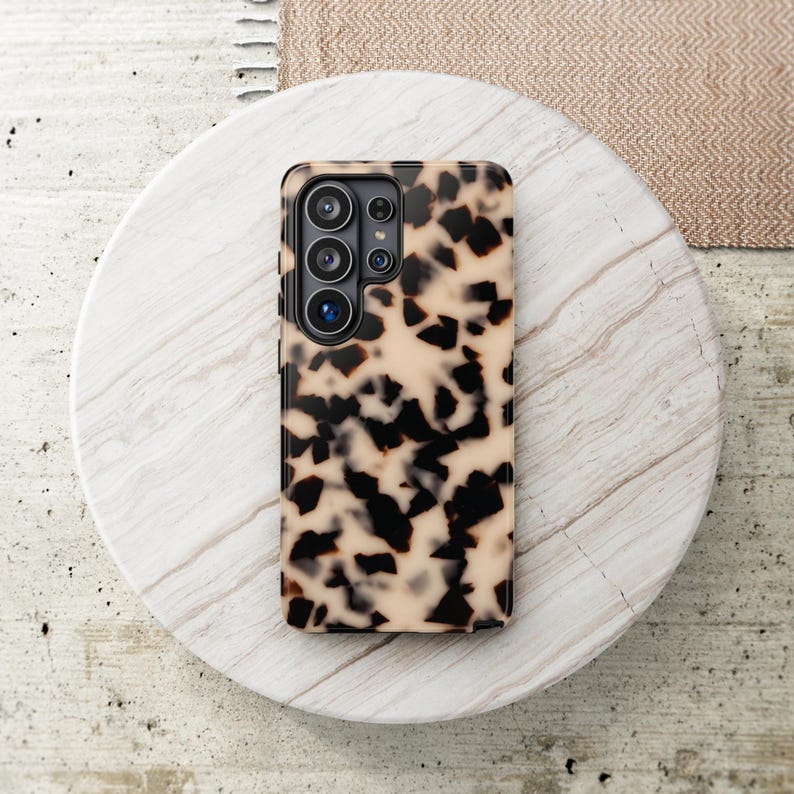 May include: A rectangular smartphone case featuring a beige and black tortoiseshell pattern. The case has a dedicated cutout section for a multi-lens camera array, showing four circular lenses and a flash component on the upper left side.