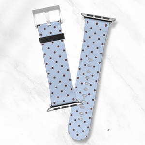 May include: Light blue Apple Watch band with a repeating pattern of brown polka dots. The band has a silver buckle and connectors. The watch band is made of a soft material.