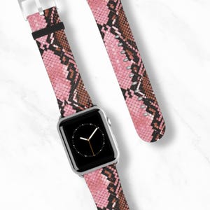 May include: A silver smartwatch with a black screen and a pink snakeskin patterned strap. The strap has brown and black accents. The watch strap is next to a matching strap. The background is white.