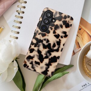 May include: A rectangular smartphone case featuring a glossy tortoiseshell pattern with irregular black and dark brown spots on a cream-colored background. The case has a cutout for a triple-lens camera system with black circular bezels.