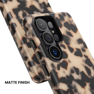 May include: A phone case with a beige and black tortoiseshell pattern and a matte finish. The case features a raised black camera cutout with multiple circular lens openings and side buttons. The text MATTE FINISH is visible.