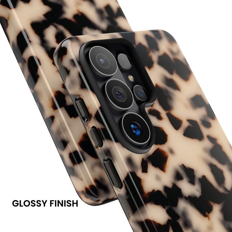 May include: A smartphone case with a glossy finish and a beige and black tortoiseshell pattern. The case features a black camera cutout frame with circular lens openings and side buttons. The text GLOSSY FINISH is printed in black.