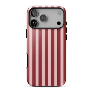 May include: A smartphone case featuring a vertical striped pattern in alternating shades of dark red and light pink. The upper section includes a rectangular camera module with three circular lenses and a flash, set against a light gray background.