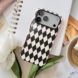 May include: A smartphone case featuring a repeating black and cream-colored diamond harlequin pattern. The case has a rectangular camera cutout at the top, revealing three circular camera lenses and a flash, with a sleek, glossy finish.