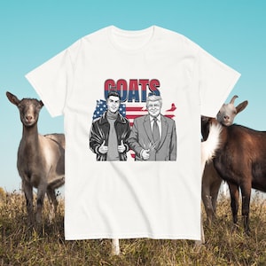 May include: White t-shirt with the word "GOATS" in red and blue lettering. The shirt features illustrations of two men giving a thumbs-up, set against a backdrop of the American flag. Two goats stand on either side of the shirt.