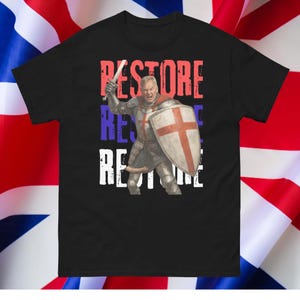 May include: Black t-shirt featuring a graphic of a knight in armor holding a sword and shield with a red cross. The word "RESTORE" is printed vertically in red, blue, and white.