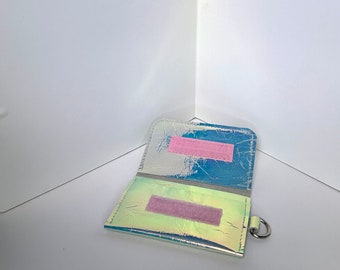 May include: A small iridescent wallet with a pink velcro closure and a silver ring. The wallet is open and the inside is visible.