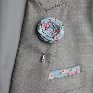 以下が含まれることがあります： グレーのスーツジャケットに、ピンクの花柄のアクセントが付いた薄いブルーの布の花がピンで留められています。花は銀色のピンで留められています。ジャケットのポケットには、それに合わせたポケットチーフが見えます。