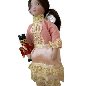 May include: A porcelain doll in a pink dress with lace trim, holding a miniature toy soldier. The doll has brown hair and is wearing a dress with a high collar and a gold-coloured belt. The toy soldier is red, black, and white.