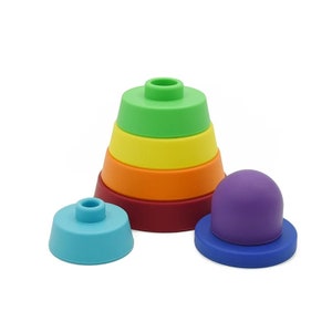 Puede incluir: Un juguete de apilar de silicona de colores arcoíris. El juguete está formado por seis piezas, cada una de un color diferente: verde, amarillo, naranja, rojo, morado y azul. Las piezas tienen forma de cono y se pueden apilar unas sobre otras.