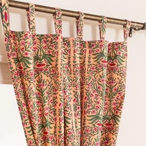 Könnte beinhalten: Pfirsichfarbene Blumenvorhänge mit einem sich wiederholenden Muster aus rosa Blüten und grünen Blättern. Die Vorhänge hängen an einer bronzefarbenen Stange mit dekorativen Endstücken. Der Stoff ist in weichen Falten drapiert.