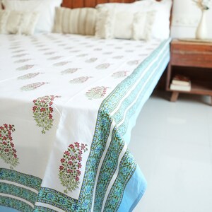 May include: White bedsheet with a floral pattern in pink and green. The border is a light blue with a decorative design. The bedsheet is on a wooden bed frame with white pillows.