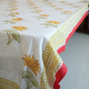 May include: White tablecloth with a repeating pattern of yellow lotus flowers and green leaves. The border features a yellow geometric design and a red edge. The cloth is draped over a table.