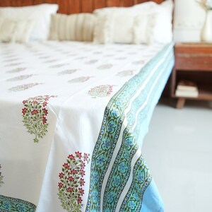 May include: White bed sheet featuring a floral pattern in pink and green. The sheet has a blue border with a repeating floral design. The bed is made up with white pillows and a wooden headboard. The overall theme is home decor.