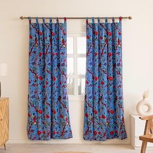 May include: Two blue floral print curtains with red flowers and birds hang in front of a window. The curtains are suspended from a bronze-colored rod. A round, woven rug sits on the floor.