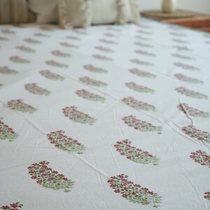 May include: White bedsheet featuring a repeating pattern of pink floral clusters and green leaves. The bedsheet is on a bed with white pillows and a wooden headboard. The overall aesthetic is soft and inviting.