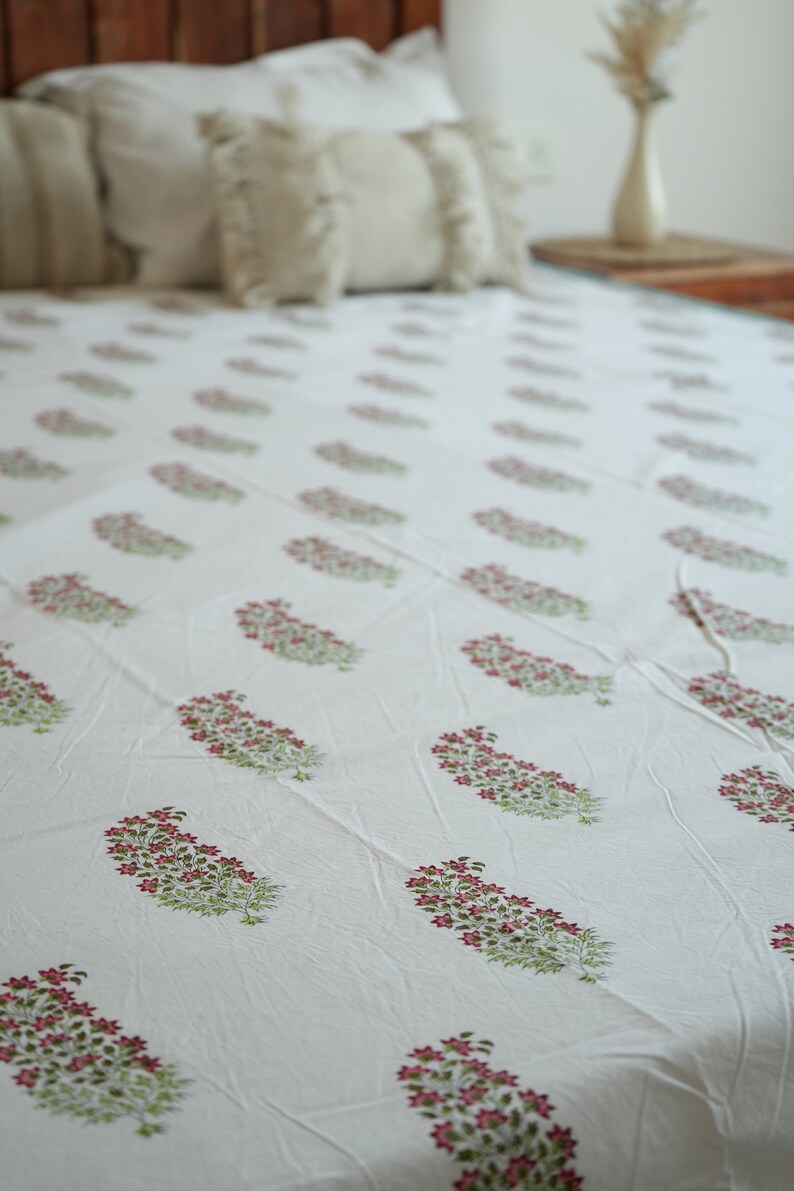 May include: White bedsheet featuring a repeating pattern of pink floral clusters and green leaves. The bedsheet is on a bed with white pillows and a wooden headboard. The overall aesthetic is soft and inviting.