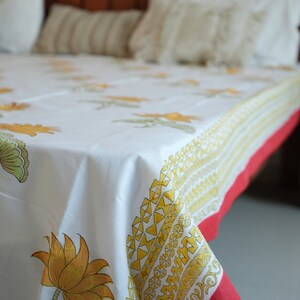 May include: White floral print bedspread with yellow and green lotus flowers and a red border. The bedspread is on a bed with white pillows and a wooden headboard. The floral pattern is repeated across the fabric.