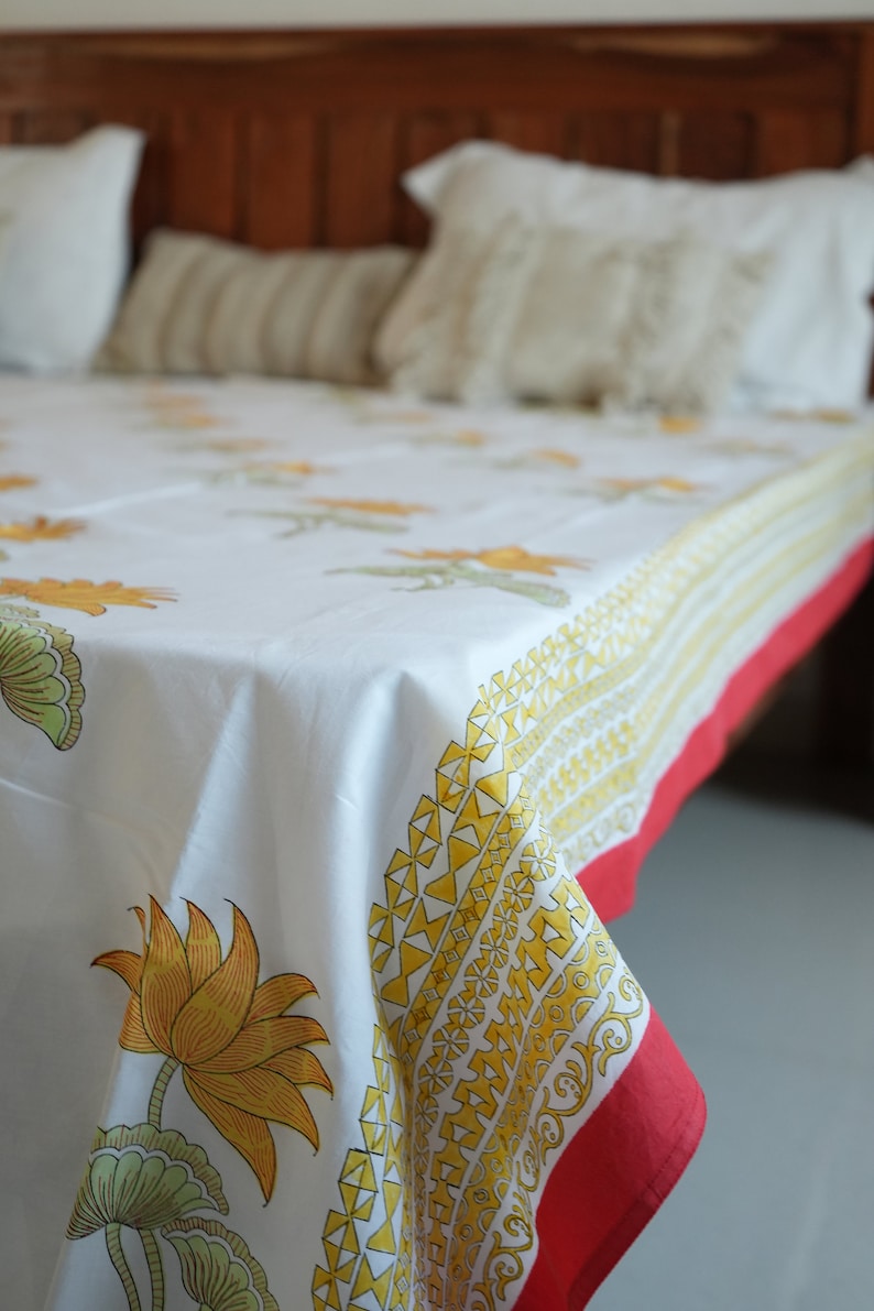 May include: White floral print bedspread with yellow and green lotus flowers and a red border. The bedspread is on a bed with white pillows and a wooden headboard. The floral pattern is repeated across the fabric.
