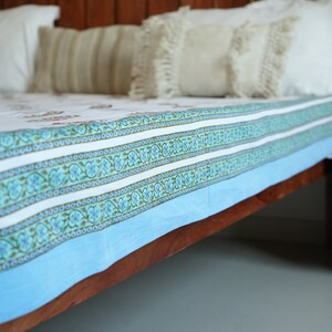 May include: A bed featuring a wooden headboard and a bedspread with a white and blue floral design. The bedspread has a light blue border and decorative patterns. Several pillows with fringe and striped patterns are visible.