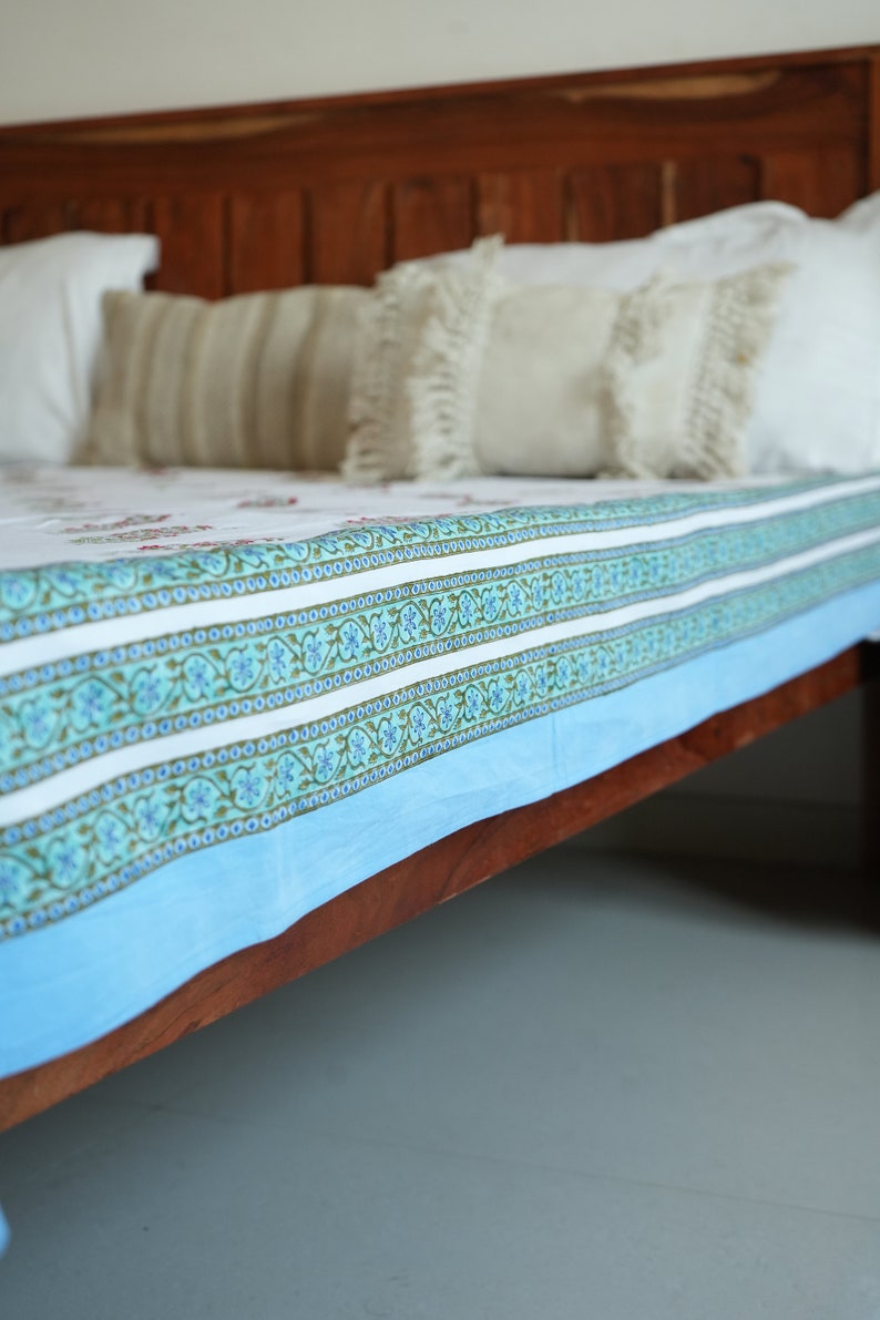 May include: A bed featuring a wooden headboard and a bedspread with a white and blue floral design. The bedspread has a light blue border and decorative patterns. Several pillows with fringe and striped patterns are visible.