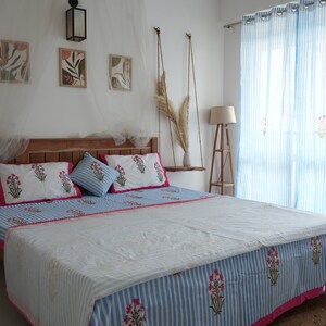 May include: A bedroom scene featuring a wooden bed frame with a blue and white striped bedspread and matching pillows. The bedding has a floral pattern with pink accents. Light blue striped curtains and framed artwork complete the room's decor.