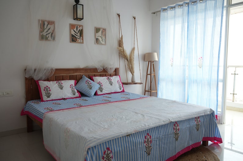 May include: A bedroom scene featuring a wooden bed frame with a blue and white striped bedspread and matching pillows. The bedding has a floral pattern with pink accents. Light blue striped curtains and framed artwork complete the room's decor.