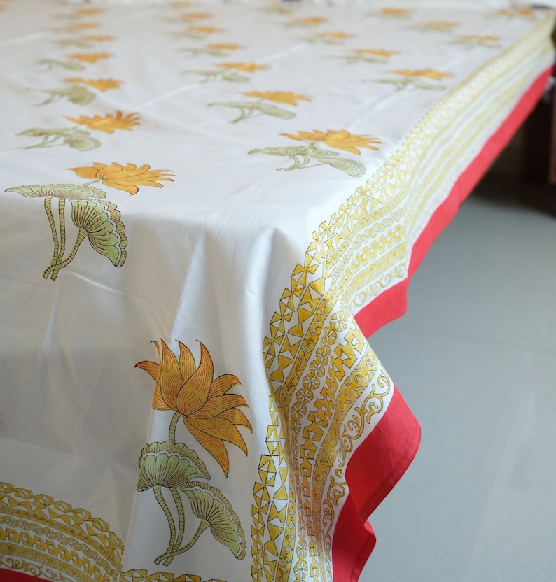 May include: White tablecloth with a repeating pattern of yellow lotus flowers and green leaves. The border features a yellow geometric design and a red edge. The cloth is draped over a table.