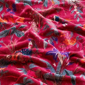 Könnte beinhalten: Nahaufnahme eines leuchtend roten Stoffes mit tropischem Blumenmuster. Das Design zeigt detaillierte Illustrationen von Blumen, Palmenblättern und Vögeln in Blau-, Lila-, Orange- und Brauntönen. Der Stoff ist stark strukturiert und drapiert.