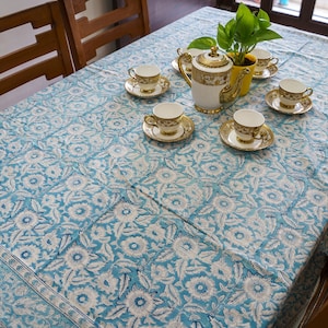 Puede incluir: Un mantel con estampado floral azul y blanco cubre una mesa de madera, con un juego de té. El juego de té incluye una tetera, tazas y platillos blancos y dorados. Una pequeña maceta amarilla con plantas verdes está en el centro.