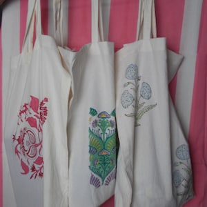 以下が含まれることがあります： ピンク、緑、青の花柄がプリントされた、オフホワイトのキャンバス地のトートバッグ3つ。バッグは、ピンクと白のストライプの背景に、金属製のロッドから吊り下げられています。各バッグには、異なる植物のプリントが施されています。