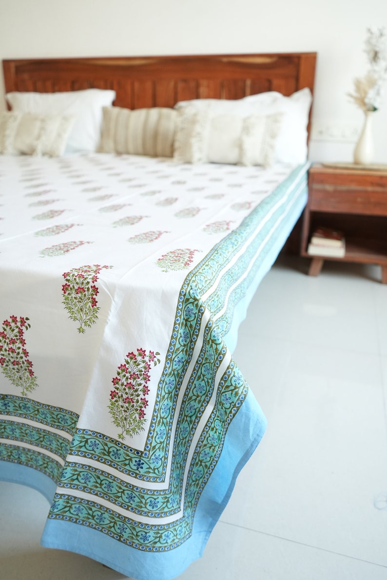 May include: White bedsheet with a floral pattern in pink and green. The border is a light blue with a decorative design. The bedsheet is on a wooden bed frame with white pillows.