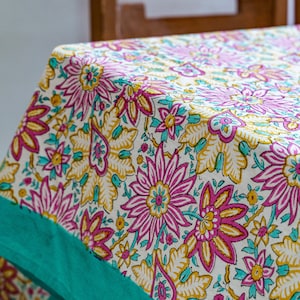Puede incluir: Un mantel floral con fondo crema y un estampado repetido de flores y hojas rosas, amarillas y turquesas. El borde es de color turquesa liso. El mantel está hecho de una tela suave.