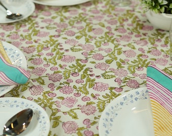 May include: A table setting with a floral tablecloth featuring pink flowers and green leaves. White plates with blue floral trim, silverware, and folded napkins in yellow, pink, and teal are arranged on the table. A small potted plant and wine glasses are also present.