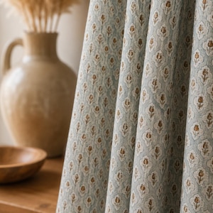 May include: A light blue curtain panel featuring a repeating pattern of small, brown, tulip-like floral motifs enclosed within white, scalloped, shield-shaped outlines. The fabric is gathered into vertical folds, displaying a textured weave.