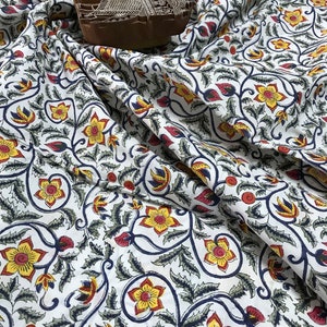 May include: White fabric with a repeating floral pattern in yellow, red, and blue. A wooden block with an intricate design rests on the fabric. The fabric is draped, showing the pattern's flow and detail. Ideal for textiles and crafts.