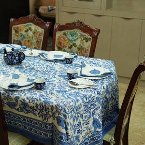 May include: A dining table set with a blue and white floral tablecloth, matching napkins, and tableware. The table is set with plates, cups, and a teapot. Wooden chairs with floral upholstered backs surround the table.