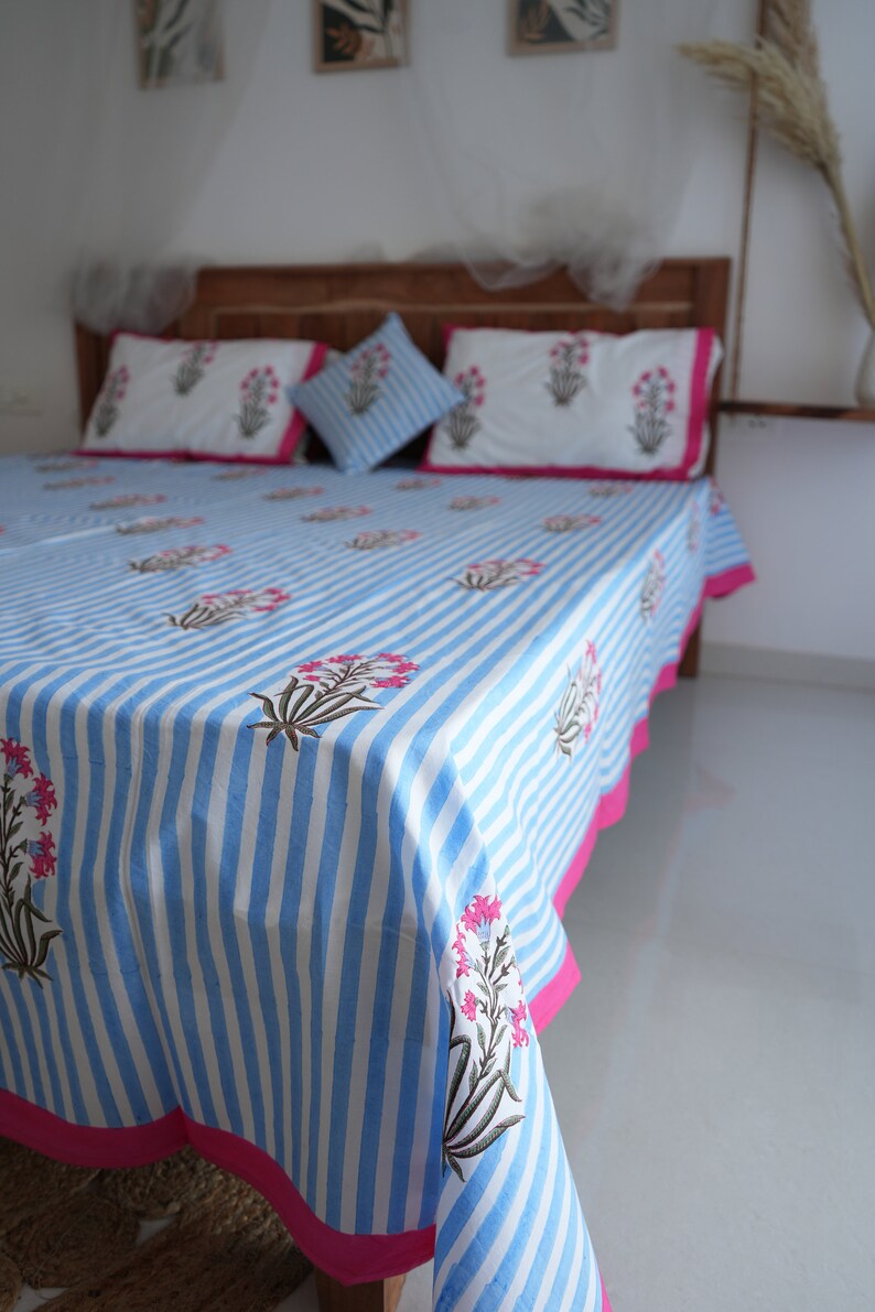 May include: A blue and white striped bedspread with matching pillowcases and a decorative pillow. The bedding features a floral pattern in pink and green. The bedspread has a bright pink border. The bed is made of wood.