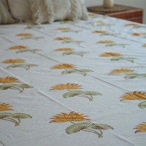 May include: White bedsheet with a repeating pattern of orange and green lotus flowers. The bed is made up with white pillows and decorative cushions. A wooden headboard and side table are visible in the background.