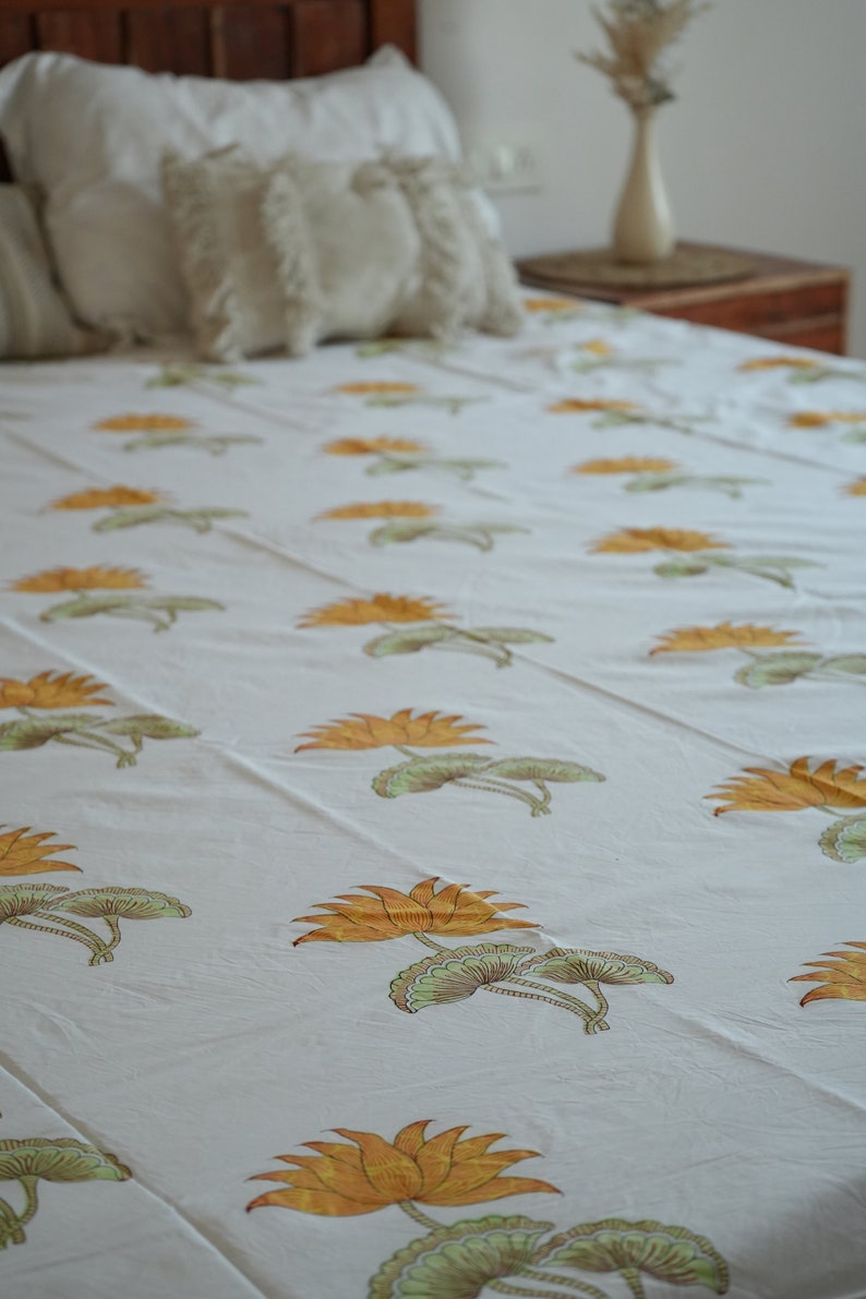 May include: White bedsheet with a repeating pattern of orange and green lotus flowers. The bed is made up with white pillows and decorative cushions. A wooden headboard and side table are visible in the background.