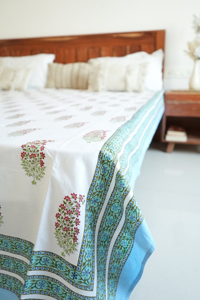 May include: White bed sheet featuring a floral pattern in pink and green. The sheet has a blue border with a repeating floral design. The bed is made up with white pillows and a wooden headboard. The overall theme is home decor.