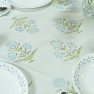 Puede incluir: Mantel blanco con un estampado floral en azul claro y verde. La mesa está puesta con platos blancos con un borde floral azul, cubiertos y una taza a juego. Un jarrón con flores está en el fondo.