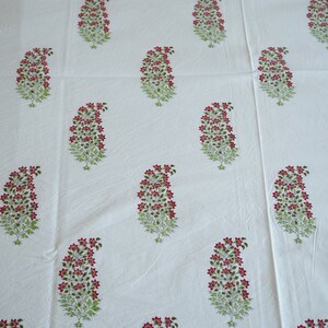 May include: White fabric with a repeating pattern of paisley-shaped floral designs. Each design features a cluster of small red flowers atop green leafy stems. The fabric appears to be cotton or linen.