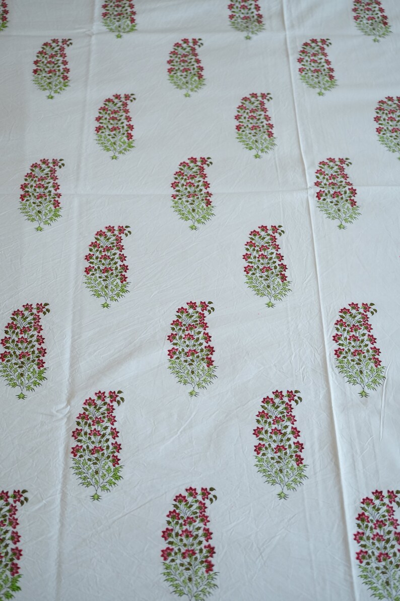 May include: White fabric with a repeating pattern of paisley-shaped floral designs. Each design features a cluster of small red flowers atop green leafy stems. The fabric appears to be cotton or linen.