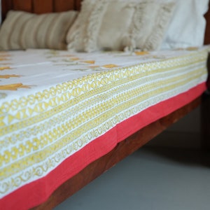 May include: A white bed sheet with a yellow floral pattern and geometric border, trimmed with a red edge. The sheet is on a wooden bed frame, with pillows in the background.