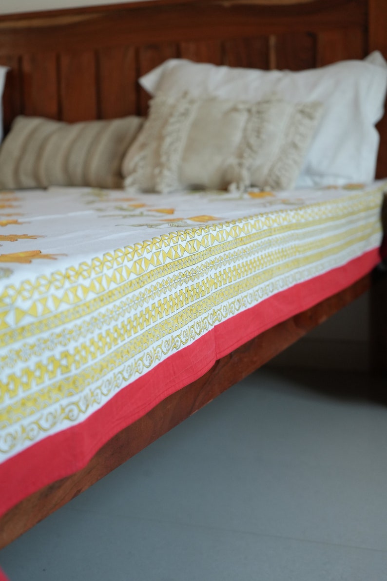 May include: A white bed sheet with a yellow floral pattern and geometric border, trimmed with a red edge. The sheet is on a wooden bed frame, with pillows in the background.